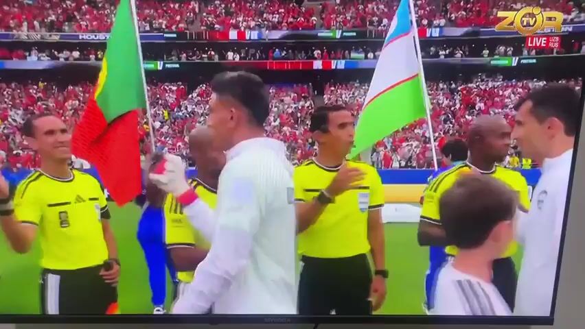 This media file captures a moment before a football match, likely during the pre-game ceremony. The scene is a stadium filled with spectators, indicated by the tiered seating in the background and a large crowd. The primary focus is on a group of individuals on the field, including players and referees, who appear to be lining up.

Several people are visible, dressed in football kits and referee uniforms. Two individuals in the foreground, one in a white long-sleeved jersey and another in a yellow and black referee kit, are shaking hands. Behind them, another person in a similar white jersey is walking away, and a player in a teal jersey, who resembles Cristiano Ronaldo, is visible in the middle ground, holding a flag. A young girl in a blue uniform stands between some of the players. Two flags are prominently displayed: one is the flag of Portugal and the other is the flag of Uzbekistan. The atmosphere seems formal and ceremonial, suggested by the handshake and the presence of flags.

The setting is a football field, identifiable by the green grass and the stadium infrastructure. The lighting suggests it could be daytime, as there are no indications of artificial stadium lights being the primary source of illumination, and no harsh shadows are cast.

Visible text includes a "ZOR TV LIVE UZB RUS" logo in the upper right corner, indicating the broadcast channel and potentially the regions where the match is being shown. The word "HOUSTON" is visible on banners in the background, suggesting a possible location, though this conflicts with the provided location context of Mashrab, Uzbekistan. There are also "FIFA" logos on the referee's uniforms. The numbers "5" and "3" are visible on the back of some players' jerseys. The overall mood appears to be professional and anticipatory, as is typical before a sporting event.