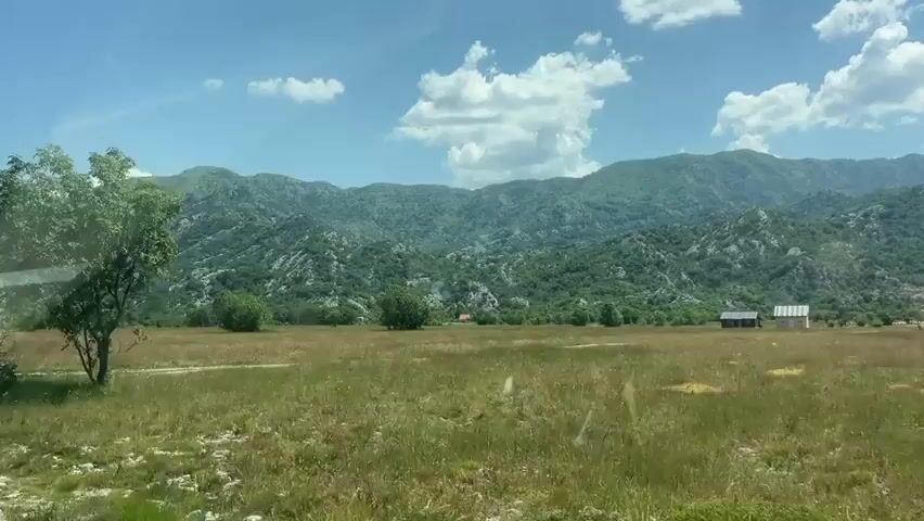 The video shows a landscape with mountains, fields, trees, and clouds. The camera appears to be moving, possibly from a vehicle, as evidenced by the slight blur and the way the scenery shifts. In the foreground, there are grassy fields with some trees. In the distance, there are rolling hills covered in greenery and rocks, forming a mountain range. The sky is blue with scattered white clouds. On the right side of the frame, a small, weathered building with a pitched roof can be seen in the distance. The overall scene depicts a rural or natural environment on a sunny day.
