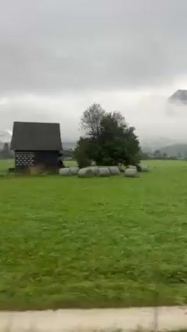 The camera pans across a large green field under a gray, overcast sky. In the distance, a small village is visible, partially obscured by fog. A mountain can also be seen in the background, its peak shrouded in mist. In the foreground, a dirt path runs alongside the field.