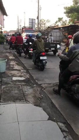 A motorcycle convoy moves down a paved road with a sidewalk. The lead motorcycle has a dashboard with a speedometer and fuel gauge. Several other motorcycles are in formation behind it. Other vehicles, including cars and a utility truck with the brand "VIAR" visible, are also on the road. Buildings and vegetation line the street. The sky is overcast.