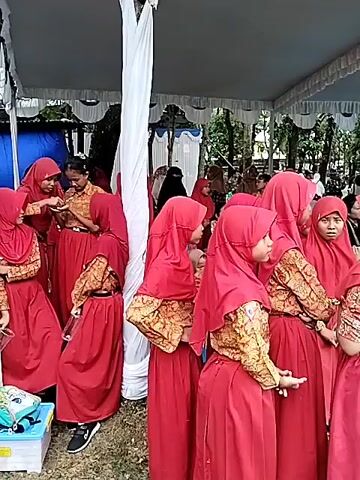The video shows a group of young girls dressed in red skirts, patterned yellow-brown tops, and red hijabs standing under a white tent. Some of them are holding small clear plastic cups. A man in a tan shirt and a woman in a hijab are visible among the girls. The camera pans to show more of the surroundings, revealing a grassy area with trees. Some adults are standing around, including a woman holding a baby, and a man in a purple jacket. The scene appears to be taking place outdoors, possibly at an event. There are other people in the background, as well as a blue tarp, and some parked motorcycles.