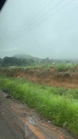 The video shows a view from a vehicle driving along a dirt road on a cloudy day. The road is wet and muddy in places, with puddles of water. To the left of the road, there is a steep bank covered in lush green grass. Beyond the bank, there are some trees and vegetation, with a few small buildings and structures visible in the distance. In the middle ground, there is an area with a few trucks and an open-sided building. The sky is overcast, and the overall impression is of a rural landscape during or after rainfall.