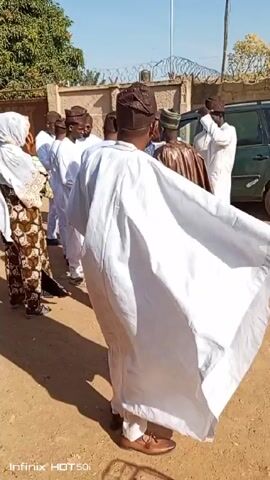 A group of people, mostly men dressed in traditional West African attire, are standing and interacting outdoors on a sunny day. The men are wearing various styles of white and brown robes and caps, and a woman in a patterned brown dress and white headscarf is also present. They are gathered on a paved area next to a low wall topped with barbed wire. A large green tree is visible in the background, along with a utility pole and a car. The individuals appear to be engaged in conversation and handshakes. The video is shot from within the group, moving slightly as it records.
