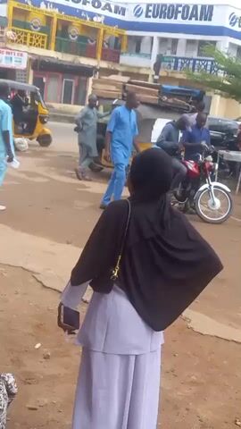 The video shows a person in a long light-colored dress and a black headscarf standing on a dirt road. The person is holding a phone in their left hand and looking towards a street scene. The street scene includes a yellow three-wheeled vehicle with goods strapped to the roof, several men in various clothing, a red motorcycle, and parked cars. A building with "EUROFOAM" in large blue letters on its façade is visible in the background.