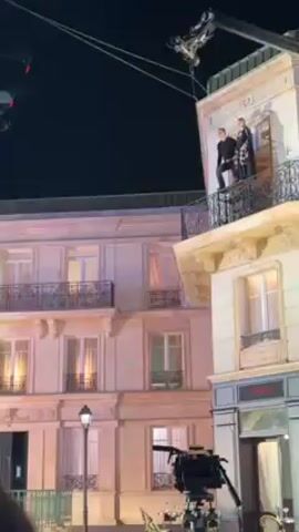 The video shows a filming scene with two figures suspended in the air on wires against a backdrop of a building facade at night. One figure, clad in a dark outfit, dangles freely while the other, adorned in a patterned outfit, seems to be grabbing onto their arm. Both are suspended by wires connected to a crane above. In the foreground, an individual in a dark jacket and beanie can be seen partially, along with filming equipment. The backdrop features a building with balconies, windows, and architectural details, with a store sign visible on the ground floor. The scene is illuminated by artificial lights, and several crew members appear to be present.