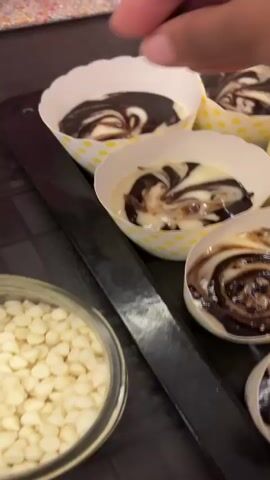A close-up shot shows a person's hand sprinkling what appear to be chocolate chips onto unbaked cupcakes. The cupcakes are in white liners with yellow polka dots, arranged on a black tray. The cupcake batter is a marbled swirl of white and dark batter, with dark chocolate sauce drizzled on top. In the background, a clear container filled with white chocolate chips is visible. The lighting suggests an indoor environment, likely a kitchen.