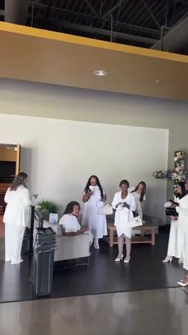 The video captures a group of women, all dressed in white, gathered in what appears to be a church lobby. One woman sits in a gray armchair, while others stand around, some holding phones or small bags. A large cross adorned with flowers stands against a white wall in the background. The environment includes modern furniture, indoor plants, and a dark polished floor, suggesting a contemporary church design. The women seem to be engaged in conversation or preparing for an event.
