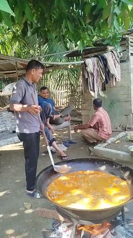 A man in a grey shirt and black pants stirs a large pot of yellow curry with a ladle. He is standing over a fire that is burning under the pot. Two other men are sitting in the background. One man, wearing a blue shirt and jeans, is sitting on a wooden bench, leaning back on his hands. The other man, wearing a red and white checkered shirt and beige pants, is crouching on the ground to the right. In the background, there is a wooden structure with clothes hanging on it and trees overhead. The scene appears to be outdoors in a rural setting.