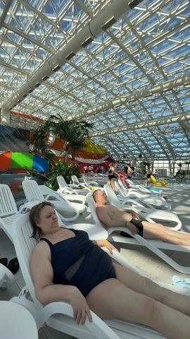 The video shows a brightly lit indoor water park. There are a few people lying on white plastic lounge chairs.  A woman in a dark swimsuit is lying on a lounge chair in the foreground with her eyes closed. To her right, a man with short brown hair is also lying on a lounge chair. He is wearing dark swim trunks and is shirtless.  In the background, other people are walking around or lounging in the water park. There are palm trees, colorful structures, and water slides. The roof of the water park is made of a grid of white beams and glass panels, allowing sunlight to filter in.  The camera is stationary.
