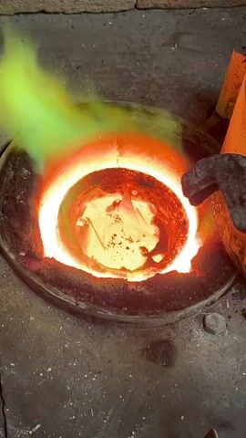 In an industrial setting, a close-up shot captures a metal smelting process within a hot crucible. A pair of long, metal tongs, heated to a glowing red at their tips, is used to feed small pieces of solid metal into a crucible brimming with incandescent molten metal, which glows a brilliant orange and yellow. Intense heat radiates from the crucible, and a vibrant green flame or smoke rises from the left side, indicating extreme temperatures and possibly the burning of impurities or specific elements within the metal. The crucible is set into a concrete or stone base, and the overall scene depicts a stage in metal preparation, likely for casting or refining. No text is visible. The location is Saint-Rémy-lès-Chevreuse, France.