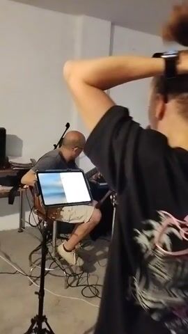 The video shows a band practicing in what appears to be a basement or a garage. In the foreground, a woman with short hair and sunglasses on her head, wearing a black t-shirt and black shorts, is adjusting her hair and singing into a microphone. Behind her, a man with a shaved head, wearing a grey t-shirt and shorts, is playing an acoustic guitar. To the right of the guitarist, another man, seen from the side, is playing a keyboard. Various musical equipment, including microphones, stands, cables, and amplifiers, are visible throughout the scene. The environment is dimly lit and cluttered, suggesting a rehearsal space.