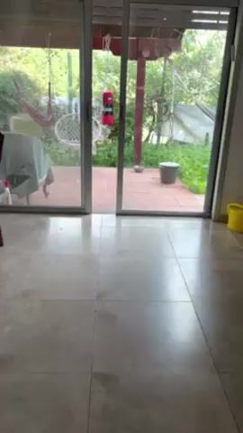 The video shows a static shot of a tiled floor in a room with a sliding glass door leading to a patio. The patio contains a table with a cover, a hanging chair, a punching bag, and various plants. The camera slowly pans left to show a wooden table and chairs inside the room. A sound similar to a child yelling is audible throughout the video.