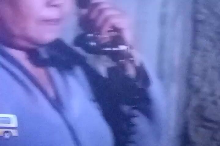 The video shows a close-up of an overweight woman with dark hair and makeup, wearing a light blue robe or blouse. She is holding a black rotary phone to her ear and speaking. The background is blurred and appears to be a wall with a textured pattern. The lighting is dim and casts a purple hue on the woman's face. The woman's expression suggests she is either surprised or displeased. The video is likely a still from a movie or television show, given the dated appearance of the phone and the overall quality of the image.