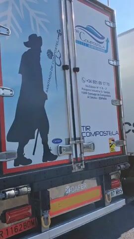 The video shows the rear doors of a refrigerated truck. The truck is white and has blue and red graphics. On the left door, there is a black silhouette of a pilgrim walking, with a staff and a scallop shell symbol. The text "SANTIAGO DE COMPOSTELA" is written vertically next to the silhouette. On the right door, there is a logo that appears to be a blue wave with an arrow. Below the logo, there is text that reads "TIR COMPOSTELA" and a phone number, "+34 881 09 17 70". There is also an address listed: "Via Edisen Nº 11 B - 2º Poligono Industrial El Tambre, 15890 Santiago de Compostela A Coruña - ESPAÑA". Underneath this, the text "TIR COMPOSTELA" is repeated. There is a yellow and black warning sticker indicating a vehicle hazard. The lower part of the truck shows red taillights and a black bumper with reflective yellow markings. The license plate on the bottom left is partially visible and reads "R 1162BDM". The video is shot from a slightly low angle, looking up at the truck doors. The lighting suggests it is daytime.