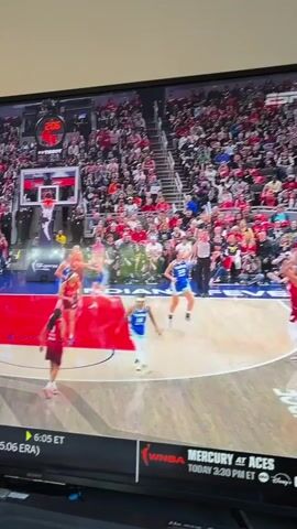 The video shows a basketball game being played on a basketball court. The teams are wearing red and blue jerseys. A player in a red jersey makes a three-point shot. A shot of the bench players in red jerseys is shown cheering. The video is being viewed on a TV. The crowd in the stands is mostly wearing red.