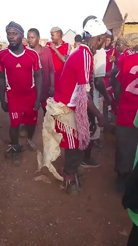 A group of people, mostly wearing red and white Adidas athletic wear, appear to be dancing or celebrating in a rural or semi-rural setting. The ground is reddish dirt. In the center is a man wearing a white cap and a red Adidas jacket with white stripes, carrying a fringed, lavender-colored item and what appears to be animal hide. Some of the red shirts have numbers on the back, such as "2" and "10". Other people around are wearing baseball caps. A man in a white shirt with the text "ZERO MALARIA – DRAW THE LINE AGAINST MALARIA" is seen. The mood appears lively and celebratory. In the background are simple buildings with peaked roofs.
