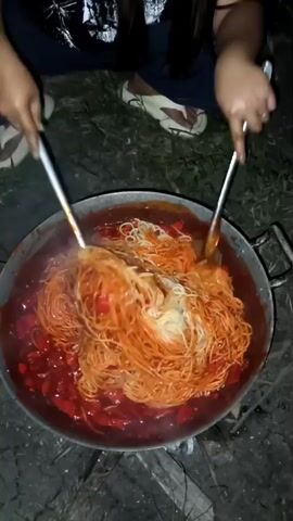 A high-angle shot shows two people cooking spaghetti in a large wok over an open fire. The wok is filled with spaghetti and a red sauce, and steam rises from the dish. The people are using large spoons to stir the spaghetti, and the flames from the fire are visible beneath the wok. The environment appears to be outdoors, with grass and dirt visible in the background. The people are wearing casual clothing, and one person is wearing sandals. The overall scene depicts a rustic and communal cooking experience.