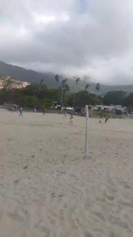 This video is taken from a beach, looking towards the water. The sand is light brown and has footprints. In the distance, there are people playing volleyball. There are also palm trees and bushes. The sky is cloudy and overcast, with mountains in the background. The video is shaky and appears to be handheld. There are no clear audio cues, only what sounds like ambient noise.