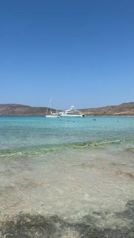 The video shows a clear blue sky over a turquoise ocean with gentle waves lapping the shore. In the distance, several large white yachts are anchored in the water. Some people are visible swimming and enjoying the ocean. The shore appears to be sandy and the water is shallow near the beach. The scene suggests a beautiful and serene day at the beach.