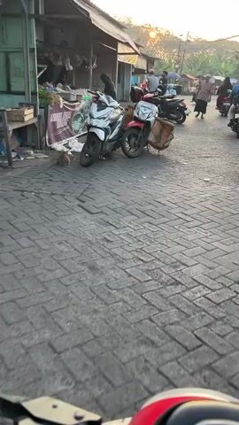 The video shows a street scene with several motorbikes and people. In the foreground, two motorbikes are parked side-by-side. The motorbike on the left is white and silver, and the one on the right is red. Behind them, more motorbikes and people are visible. The setting appears to be a marketplace or a busy street with shops on the side. The ground is paved with interlocking bricks. Sunlight is visible in the background, suggesting it might be morning or late afternoon. A person on a light blue motorbike is seen passing by, carrying a crate on the back. Another person in a brown robe and a white cap is walking in the street, looking towards the camera.