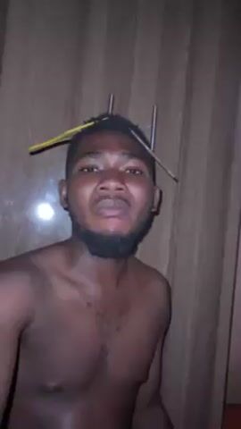 A shirtless man with a beard stands in front of a wooden closet. Two metal hangers are stuck in his hair. He exhales smoke into the camera, wide-eyed. He then holds a cigarette in his right hand and smiles.