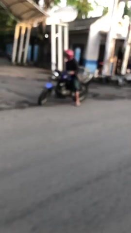 The video shows a person on a motorcycle in a blurry outdoor setting. The person is wearing a pink helmet and appears to be stationary on the motorcycle. The motorcycle is dark in color. The environment looks like a road or street with some structures in the background. The lighting suggests it might be dusk or dawn.