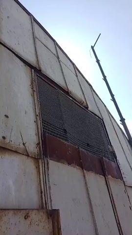 The video shows a close-up view of the exterior of a weathered, corrugated metal structure. The metal panels are a dull, off-white color with significant rust stains, particularly around a large, screened opening. The screening is a dark metal mesh. A thin, dark pole or antenna extends diagonally upwards from the top right of the frame. The lower portion of the structure appears to have a large, rectangular metal door or panel, also showing signs of rust and wear. This panel has a latch mechanism with a chain attached. The sky is visible at the top of the frame, appearing bright and possibly overcast. The camera angle is looking upwards at the structure.