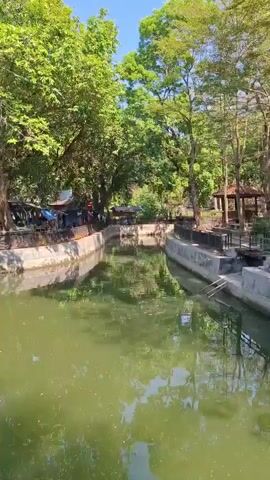 A serene river flows through a park-like setting. Lush green trees line both banks of the river, their leaves reflecting in the calm water. On one side of the river, a row of market stalls or food vendors is visible, with awnings providing shade. People are seen gathered around these stalls, suggesting a lively atmosphere. Small boats, possibly paddle boats, are present in the river, with a few in motion. The scene is bathed in sunlight, indicating a pleasant day. A black metal fence runs along the edge of the walkway beside the river, separating it from the grassy bank. The water is a murky green color, and the overall impression is one of a peaceful, natural environment with some signs of human activity.