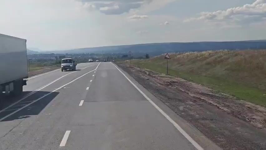 A video shows a view from inside a vehicle driving on a highway. The road is paved and has white lane markings. To the left of the road, there are grassy areas and a small town or village in the distance. Hills are visible on the horizon. To the right of the road, there is a dirt embankment with dry grass and a few trees. There are several cars and trucks on the road, all moving in the same direction as the camera. The sky is partly cloudy, with a few fluffy white clouds. There are traffic signs along the road. One sign indicates a speed limit of 70. The video is taken during the daytime, with bright sunlight.