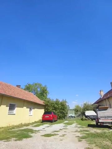 The video shows a street view on a clear day with a bright blue sky. The camera pans across a residential area, revealing houses, a garage, a street light, a trash can, and a red car. The houses are of varying architectural styles and colors, with some featuring gardens and trees. The road appears to be paved, and there are patches of grass and weeds along the sides. The video captures the general ambiance of a quiet suburban neighborhood on a sunny day.