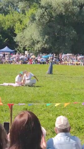 The video opens with a wide shot of an outdoor event, likely a festival or fair, with a crowd of spectators sitting on the grass in the background. Two individuals are in the foreground, positioned on the grass. One, wearing a green cap and white clothing, is seated on a pink blanket. The other, in white clothing, is crouched down next to them. In the mid-ground, a woman in a long, flowing blue dress stands and plays a violin. The environment is a grassy field with trees and tents in the background. The lighting suggests it is daytime with clear skies. The camera is positioned behind a few spectators, whose heads and shoulders are visible in the bottom of the frame.