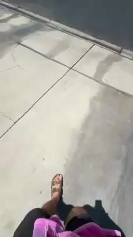 A person is walking along a cracked concrete sidewalk, wearing brown sandals and a pink towel or garment. The camera, presumably held by the person, pans up from their feet to reveal a sunny residential street in Palm Desert, United States. The street is asphalt, with well-maintained green lawns in front of single-story houses that have terracotta-colored roofs. Numerous tall palm trees line both sides of the street, and a clear blue sky with some wispy clouds is visible above. A white car is parked in a driveway. The neighborhood appears quiet and is bathed in bright daylight. No visible text is present.