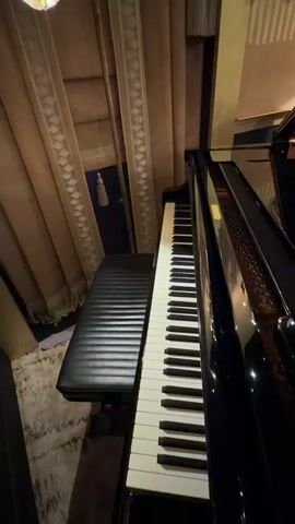The video shows a black grand piano with the lid open in a room that appears to be part of a luxury hotel or residence. The piano's interior is visible, showing the strings and soundboard. The room is decorated with patterned wallpaper, curtains, and a rug. In the background, through an open doorway, two people can be seen walking in a different room. The room appears to be luxuriously furnished with a dark wood cabinet, decorative items, and a light-colored patterned rug. The camera moves slightly while filming, giving a sense of the room's layout.
