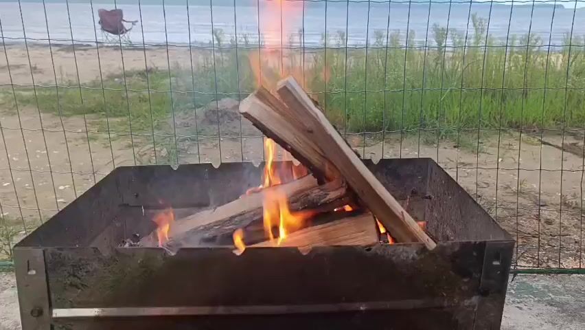 A fire burns in a metal barbecue grill. The grill contains several logs of wood, which are engulfed in flames. In the background, a beach can be seen with a red chair on the sand. Beyond the beach is a body of water, and a fence encloses the area around the grill. The scene appears to be outdoors, possibly at a campsite or park.