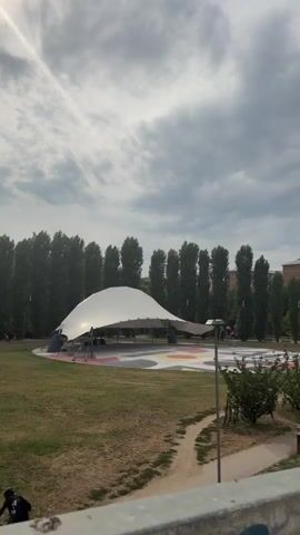 The video shows an outdoor park with a large, white, futuristic-looking tent or shelter in the center. The tent is supported by poles and has a curved, flowing shape. In front of the tent, there is a circular area on the ground with colorful geometric patterns. The park has grassy areas, trees, and a pathway. There are some people visible in the distance, but they are not clearly identifiable. The sky is cloudy. The video pans to the right, showing more of the park, including a concrete wall with graffiti on it, and a railing.