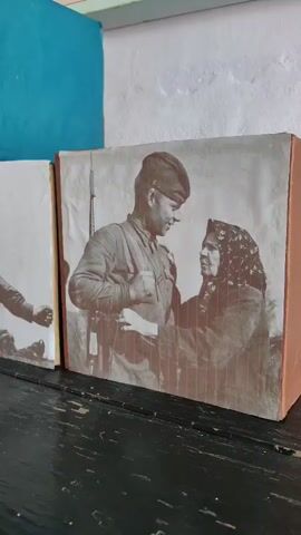 The video displays a collection of sepia-toned photographs mounted on cubic objects, possibly book covers or display blocks, arranged on a dark wooden surface. The photographs depict scenes featuring individuals in military attire, reminiscent of wartime. In the foreground, a photograph shows a soldier in uniform interacting with an older woman wearing a headscarf. To the left of this image, another photograph features a soldier's arm and hand, and further to the left, a partial view of another photograph is visible. Behind the main photograph, two other cubic objects are stacked, one with a blue side and another with a photograph of a soldier in profile. Below these, another row of cubic objects is visible, each adorned with a photograph of a soldier. One shows a soldier in uniform with medals, another shows a soldier in a dynamic pose, aiming a weapon upwards, and another appears to be a close-up of a soldier's face. The background shows a white, textured wall, with glimpses of what might be additional photographs or printed materials mounted on it. The lighting appears to be natural, casting soft shadows.