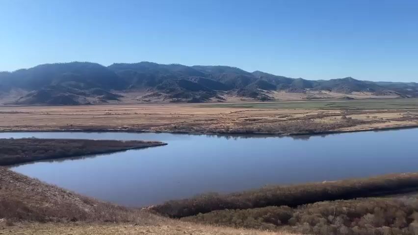 A wide river flows through a valley with mountains in the background under a clear blue sky. The valley floor consists of dry grasslands and a few patches of green. Patches of trees and shrubs line the riverbanks, with a hill in the foreground on the left. The river meanders, creating islands and inlets. The overall scene is peaceful and scenic.