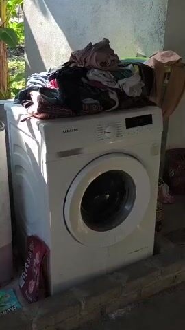 A white Samsung washing machine is shown outdoors. A pile of laundry sits on top of the machine. To the left of the washing machine is a partially visible pink and white appliance, possibly another washing machine. To the right of the washing machine, a woven basket or container is visible. The washing machine is placed in a sheltered outdoor area, possibly a patio or veranda, with a concrete floor and a textured wall behind it. Lush green foliage and a banana tree are visible in the background. The scene is illuminated by natural sunlight, casting shadows.