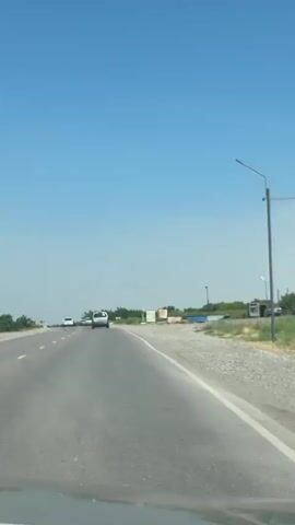 The video shows a view from inside a car traveling on a highway on a clear, sunny day. The camera is pointed forward, capturing the road ahead and the surrounding landscape. The road appears to be a two-lane highway with a white line marking the lanes. There are other vehicles on the road, including cars and possibly a truck in the distance. The landscape to the sides of the road consists of vegetation, including trees and bushes, and some signs of human activity, such as buildings or structures. The windshield of the car has some debris and a crack. The overall impression is one of a typical road trip experience.