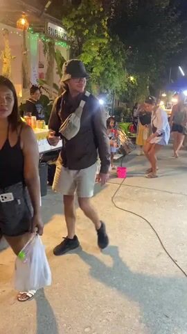The scene shows a nighttime street market with people milling around. In the foreground, a man in a bucket hat, shorts, and a fanny pack walks past. To his left, someone is holding a plastic bag. In the middle ground, several people are gathered around a vendor selling what appears to be lottery tickets. A small girl in a white dress interacts with the tickets, seemingly under the supervision of adults. There's a pink bucket nearby, possibly for collecting tickets or prizes. A blackboard displays menu items and prices. The market is lit with string lights and lanterns, creating a festive atmosphere. Trees and other foliage provide a backdrop to the scene. The ground is concrete and there is a power cord running across the scene.