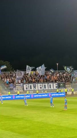 A nighttime soccer match is underway. The camera is focused on a section of stadium seating filled with fans. Many of the fans are holding up flags, primarily in blue and white, and some appear to be wearing matching team colors. A large banner is displayed horizontally across the seating area, with the word "BOJBONE" prominently written in white letters with a black outline. Below the seating area, players in blue uniforms are visible on the green soccer field. The sky above is dark, indicating it is nighttime, and the stadium lights are illuminating the field and the stands. There are also advertisements along the lower edge of the stands.