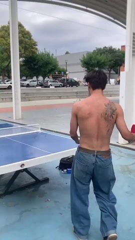 Two young men are playing table tennis outdoors. One man, wearing a white t-shirt and blue jeans, stands on the left side of the table, holding a red ping pong paddle and serving the ball. The other man, shirtless and wearing blue jeans, stands on the right side of the table, holding a pink ping pong paddle and ready to receive. The table is blue with a white net. The ground is blue. In the background, there are trees, parked cars, and buildings. The sky is overcast.