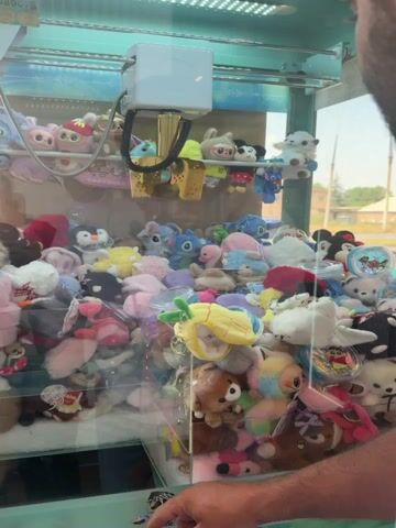 The video shows a claw machine filled with plush toys. A person's head and shoulder are visible on the right side of the frame, looking into the machine. On the left side of the frame, another person's hand is visible, appearing to be operating the controls of the machine. The plush toys are varied in color and design, with some appearing to be characters from popular media. The environment appears to be indoors, possibly an arcade or entertainment center, with bright lighting and a colorful background visible behind the toys. The claw mechanism is visible at the top of the machine, suspended over the toys. The overall impression is of someone attempting to win a prize from the claw machine.