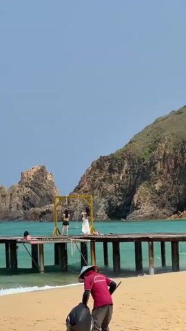 The video shows a beach scene with a long wooden pier extending into the ocean. The water is a turquoise color. Behind the pier is a rocky, mountainous island covered in greenery. On the pier, there is a yellow swing set, people posing for photos, a white piano, and a yellow telephone booth. A person in a red shirt and straw hat is sweeping the sandy beach in the foreground. A stack of yellow inner tubes are visible in the bottom right corner of the frame. The sky is a clear, bright blue.