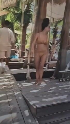 Two women in bikinis are standing on a wooden deck with palm trees and thatched roofs in the background. The woman in the foreground, on the left, is wearing a tan bikini and sunglasses. She is looking towards the left and appears to be walking. The woman behind her, on the right, is wearing a pink and orange bikini and is standing next to a wooden pillar, looking towards the front. Other people and lounge chairs are visible in the background, suggesting a beach or resort setting.