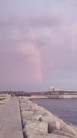 A vertical, handheld shot captures a coastal scene. The sky is filled with soft purple clouds, and a faint rainbow arcs across the upper half of the frame. Below the sky, a paved walkway runs along the left side of the image, leading away from the viewer. Several people are visible on the walkway, appearing as small figures in the distance. To the right of the walkway, a rocky embankment juts into the calm, purplish water. In the background, a long, low building with a reddish-brown roof is visible, and behind it, a tall, light-colored modern building stands against the horizon. Several birds are flying in the sky. The overall mood is tranquil and serene, with the soft colors of the sky and water creating a dreamlike atmosphere.