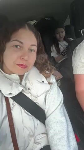 The video features a woman in the front passenger seat of a car, recording a selfie video. She has long, curly brown hair, green eyes, and freckles, and is wearing red lipstick. She is dressed in a white puffer vest over a light grey long-sleeved shirt and has her seatbelt fastened. Initially, she smiles directly at the camera, then makes a peace sign with her right hand, displaying painted nails. In the back seat, a child with dark hair, wearing a light-colored t-shirt with a graphic and striped fingerless gloves, is visible, smiling and laughing. A partial view of another person's arm or shoulder is seen on the right side of the frame. The interior of the car is visible, and music is playing in the background. The scene appears to be taking place during the daytime in Горняк, Russia, with both the woman and child appearing cheerful.