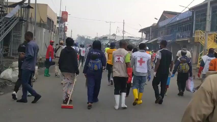 Here is a detailed description of the video:

**Scene:** The video shows a group of people walking down a street in what appears to be an urban area. The street is lined with buildings on both sides.

**People:**
*   The group consists of approximately 15-20 individuals.
*   Several people are wearing vests or jackets with the Red Cross symbol on them, indicating they may be part of a relief or aid organization.
*   Some people are wearing masks.
*   Some people are wearing boots.

**Objects:**
*   One person carries a broom.
*   Some people have backpacks.
*   Some carry plastic bags.

**Actions:**
*   The group is walking away from the camera.
*   The people seem to be moving purposefully, possibly conducting some kind of activity.

**Interactions:**
*   The group appears to be organized, suggesting they are working together.
*   There is chatter and conversation in the background.

**Environment:**
*   The environment appears to be a busy street, possibly a market area.
*   The sky is overcast.
*   The overall atmosphere seems calm, though the context suggests a possible clean-up effort or a response to an event.

Without further context, it's challenging to definitively state the purpose of the gathering, but the elements suggest a coordinated effort, possibly involving humanitarian aid, clean-up, or community support.