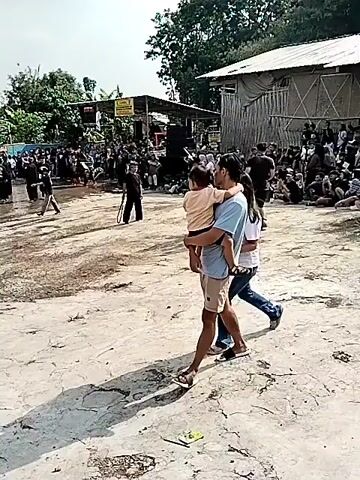 The video shows an outdoor event with a crowd of people gathered. In the foreground, a man carries a child on his shoulders while walking, accompanied by a woman. The ground is dirt and appears uneven. In the background, there is a stage with speakers and what appears to be a performance taking place. People are seated and standing around, watching the event. The overall atmosphere seems to be festive.