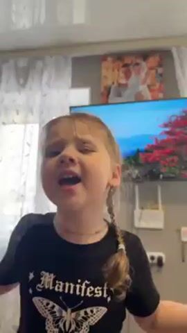 A little girl, likely between the ages of 3 and 5, is standing in what appears to be a living room. She has blonde hair styled in braids, a fair complexion, and is wearing a black T-shirt with white lettering. She looks directly at the camera, singing and holding up small objects like a makeup brush and what appears to be a green toy. Behind her, there's a window covered with a lace curtain, a television displaying a vibrant image, and a framed picture hanging on the wall. The room is illuminated by natural light.